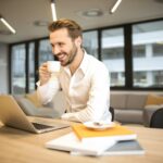 Man enjoying a coffee break while working in a modern office with laptop and books.